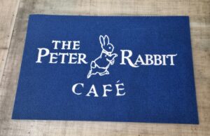 An image of the Peter Rabbit Mat. It has a dark blue background and a white outline of a rabbit and white lettering saying 'The Peter Rabbit Café'