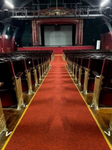 A picture of the theatre taken from the top of the aisle, showcasing Claret in action down the middle aisle, with rows of chairs on either see, and the centre stage at the end of the room